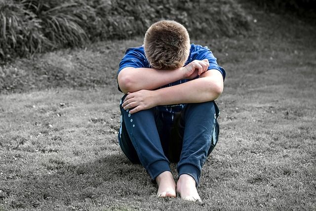 Photo of a sad boy sitting on grass with his arms resting on his knees, and his head looking down and resting on his arms
